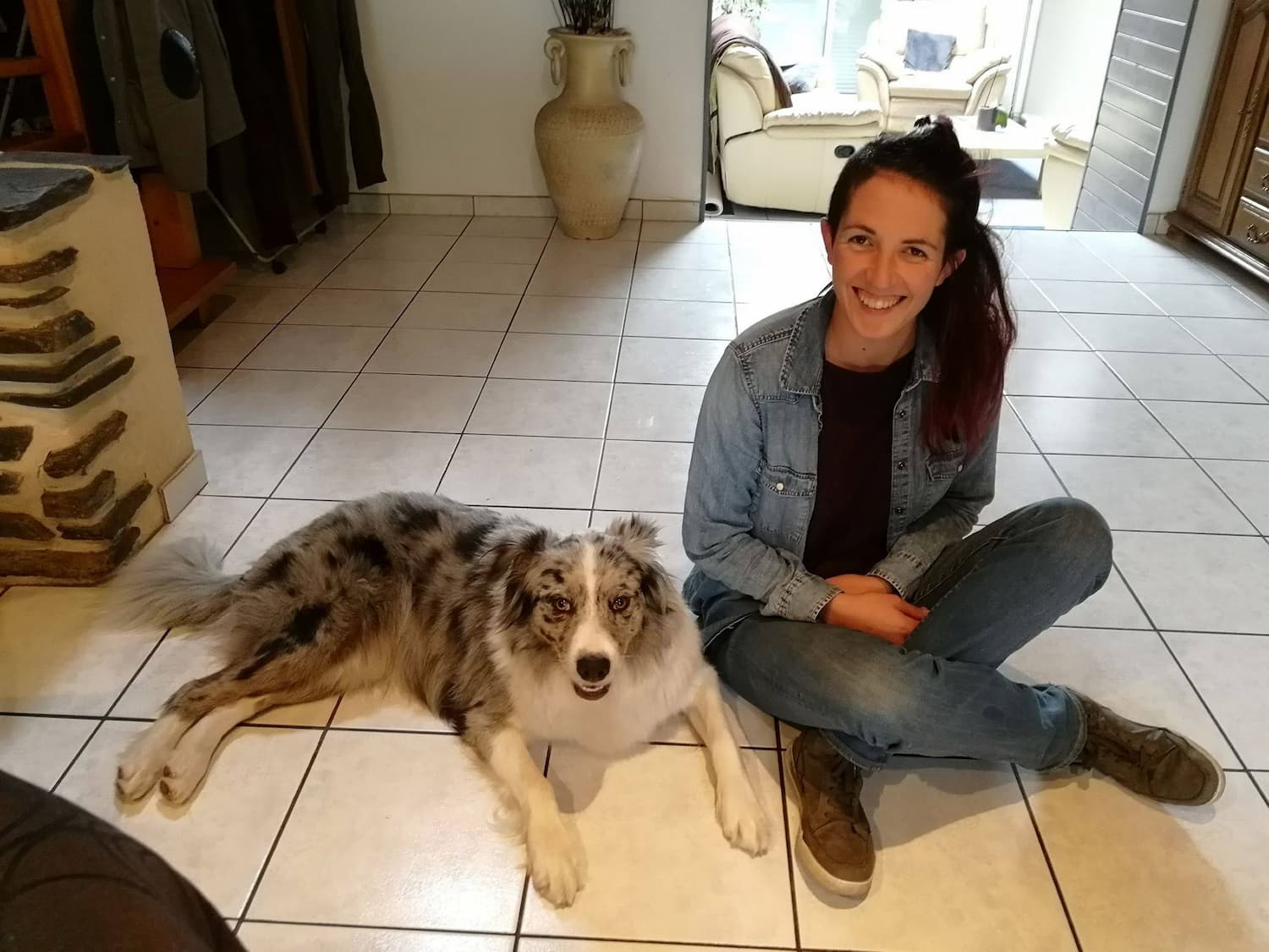Céline assise au sol à côté d'une chienne Berger Australien qu'elle a accompagné. La chienne est couchée et détendue. Elles regardent toutes les deux la caméra.